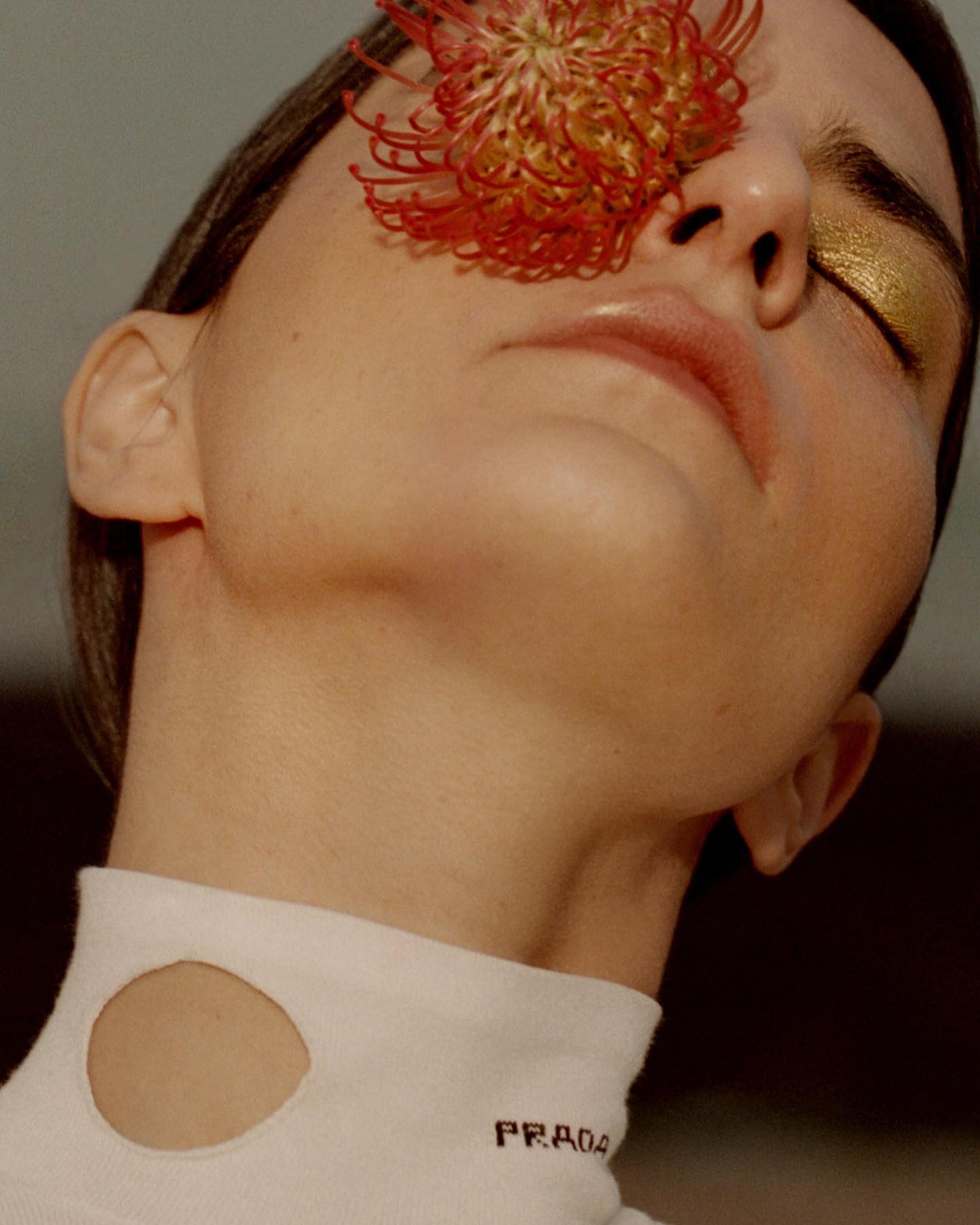 Woman with closed eyes poses with a flower on her face. She wears a white turtleneck sweater, photographed in a close-up shot.