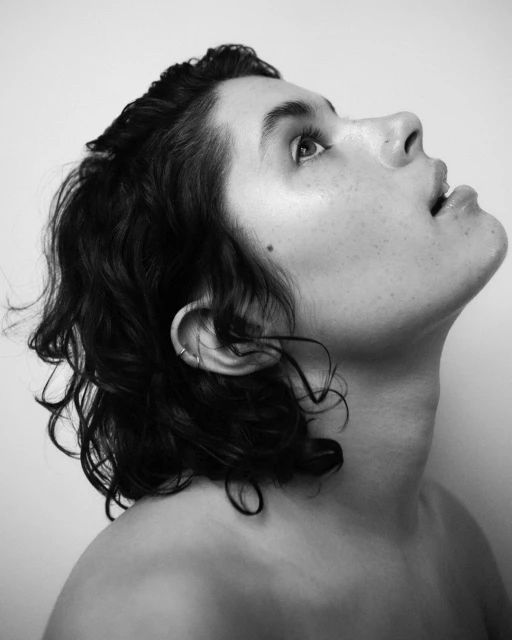 Black and white portrait of a person looking up with a tilted head, soft lighting, and curly hair, set against a plain background.