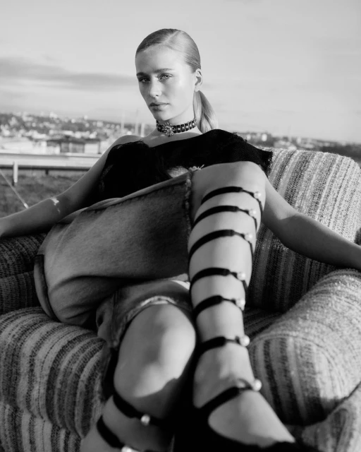Monochrome portrait of a young woman in a black top and unique strapped footwear, seated on a striped sofa with a cityscape backdrop.