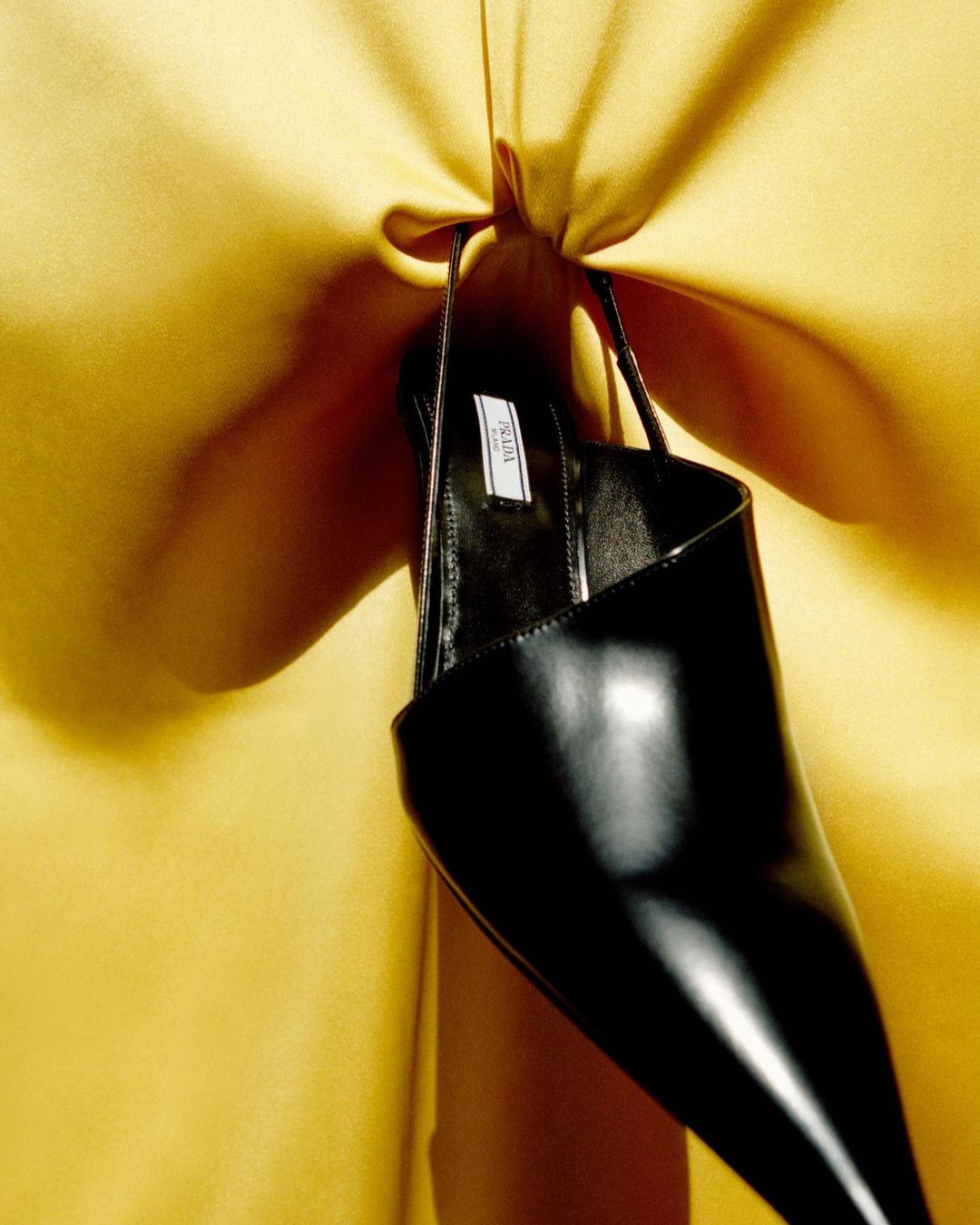 Close-up studio shot of a glossy black slingback heel by Prada against a mustard yellow draped fabric background with soft studio lighting.