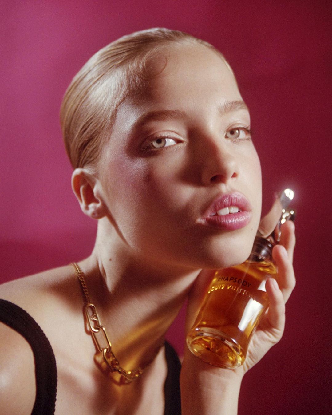 Close-up of a fair-skinned woman with a gold chain necklace holding a perfume bottle against a pink background in a beauty shot.