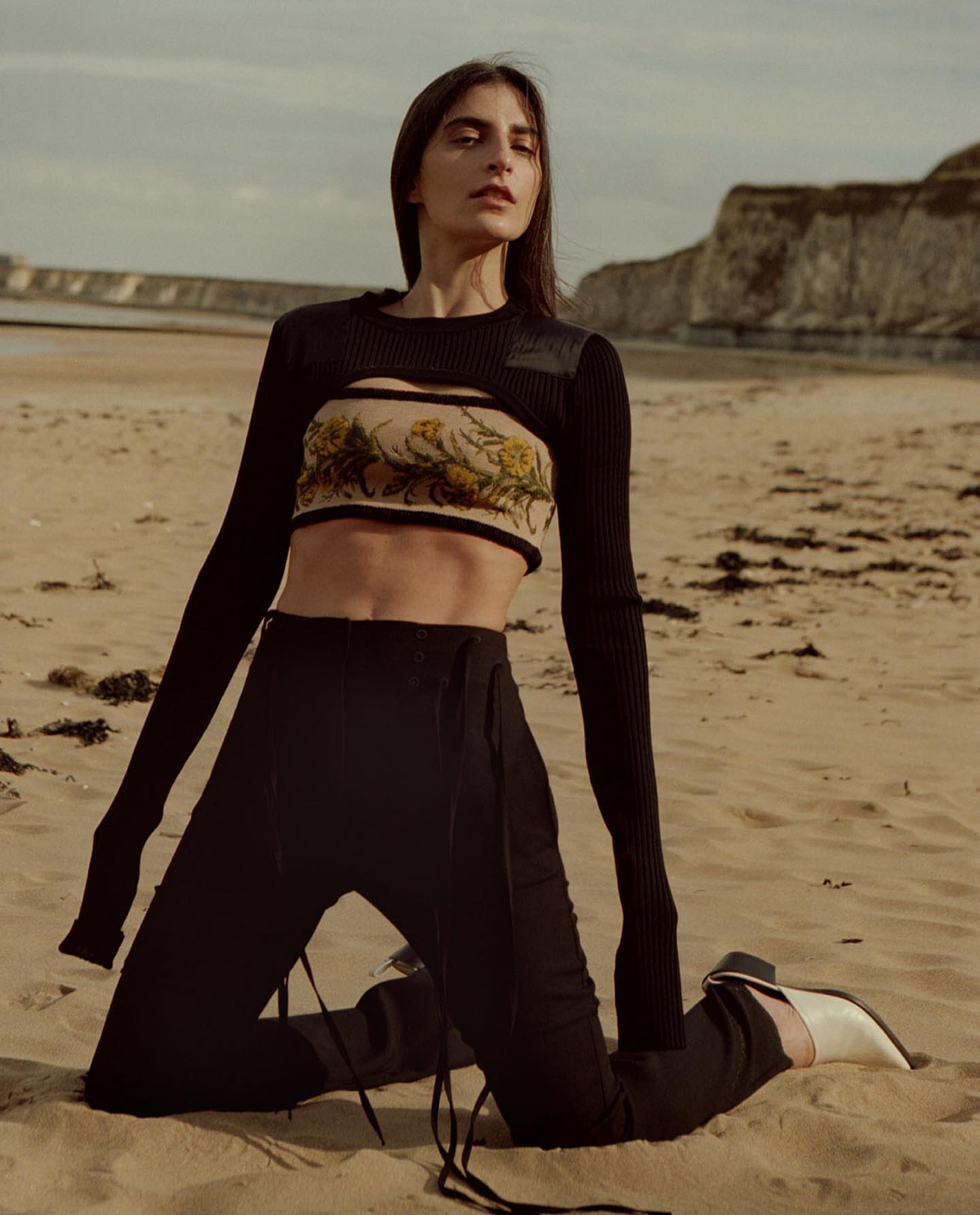 Woman kneeling on a sandy beach wearing a black knit top with a patterned band and dark trousers, photographed by Johanna Nyholm.