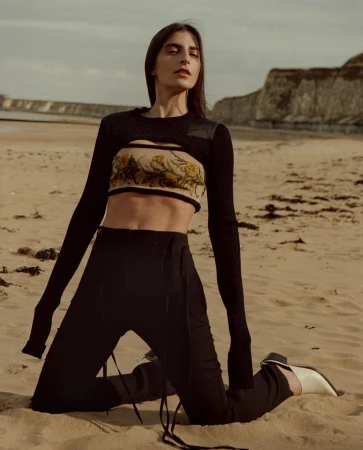 Woman kneeling on a sandy beach wearing a black knit top with a patterned band and dark trousers, photographed by Johanna Nyholm.