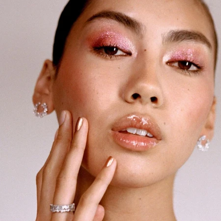 Close-up shot of a woman with pink glitter eyeshadow, glossy lips, and gold-painted nails wearing floral earrings and a diamond ring.