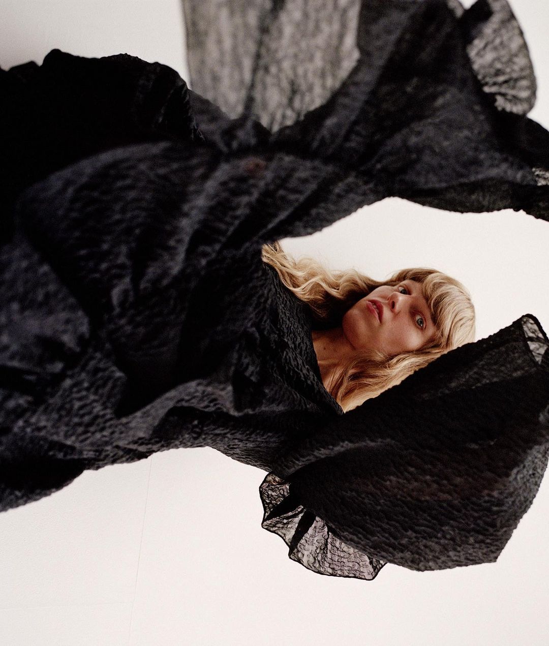 Woman with blonde wavy hair posing while covered in black textured flowing fabric against a white background in portrait format.