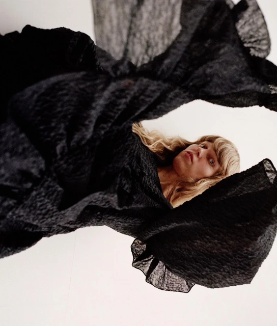 Woman with blonde wavy hair posing while covered in black textured flowing fabric against a white background in portrait format.