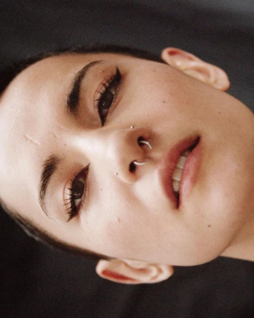 Close-up portrait of a woman with nose piercings looking up at the camera, set against a dark, plain background, beauty photography.