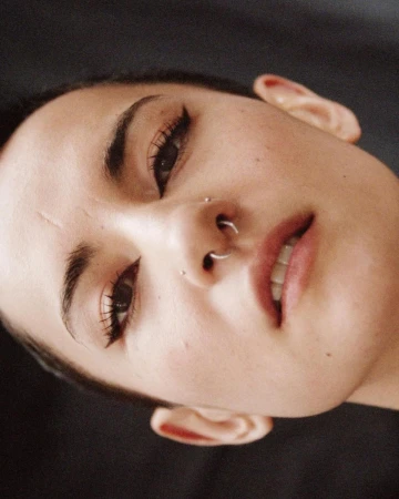 Close-up portrait of a woman with nose piercings looking up at the camera, set against a dark, plain background, beauty photography.