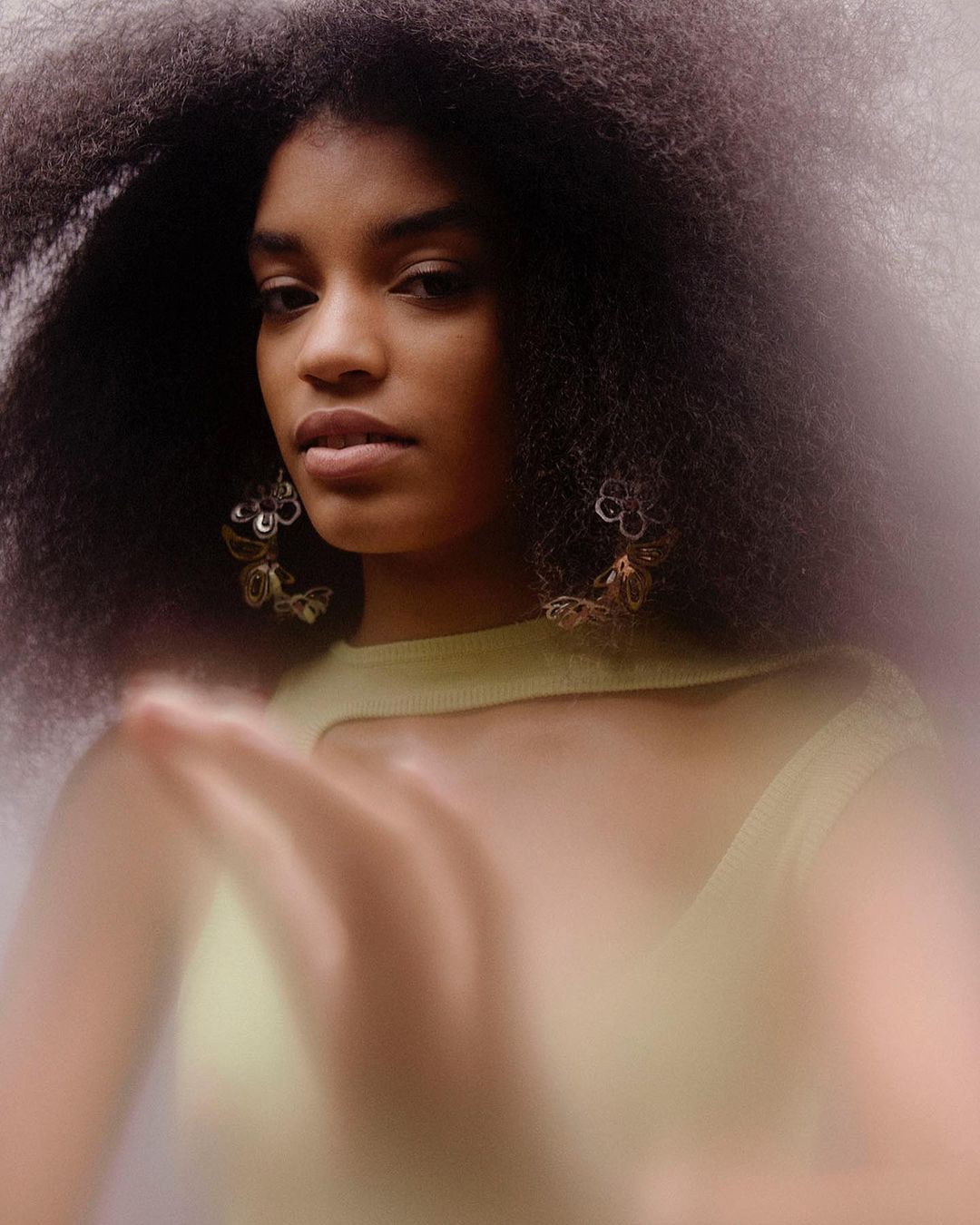 Portrait of a Black woman with natural afro hairstyle, wearing statement earrings and a light green top against a blurred background.