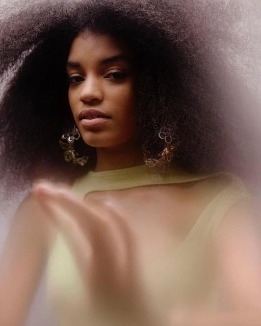 Portrait of a Black woman with natural afro hairstyle, wearing statement earrings and a light green top against a blurred background.