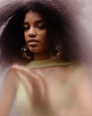 Portrait of a Black woman with natural afro hairstyle, wearing statement earrings and a light green top against a blurred background.
