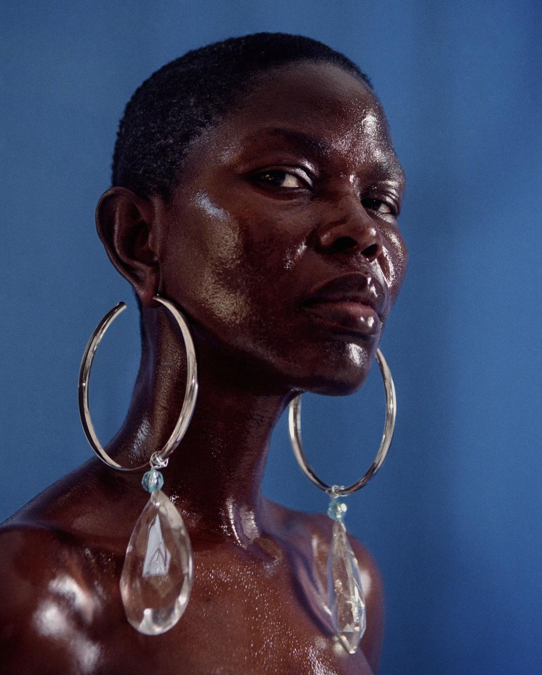 Close-up portrait of a Black woman with short hair, large hoop earrings with clear pendants, against a solid light blue background.
