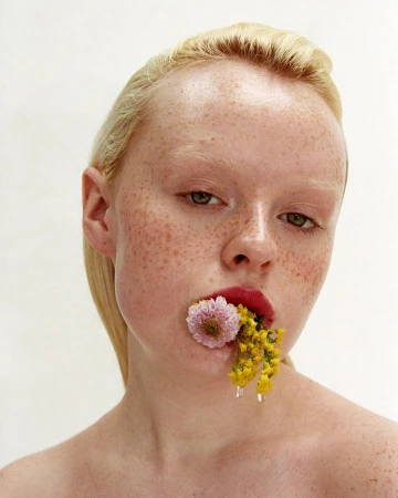 Close-up portrait of a blonde woman with freckles holding pink and yellow flowers in her mouth against a plain white background.