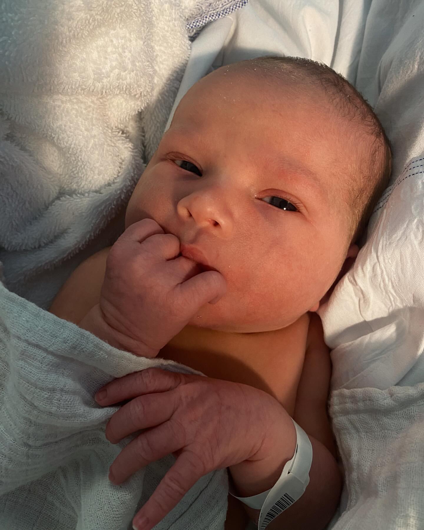 Close-up portrait of a newborn baby with fair skin wrapped in a soft white and blue blanket, with their fingers near their mouth.