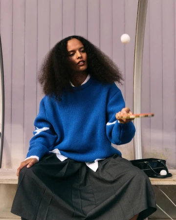 Young adult woman in a blue sweater and dark skirt plays ping pong indoors, with a ping pong paddle and a ball in mid-air.