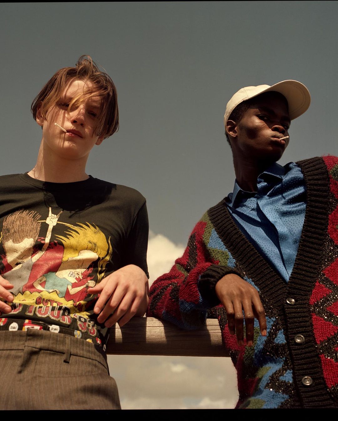 Two young adults pose outdoors on a sunny day. The individual on the left wears a graphic tee, while the person on the right sports a colorful cardigan.