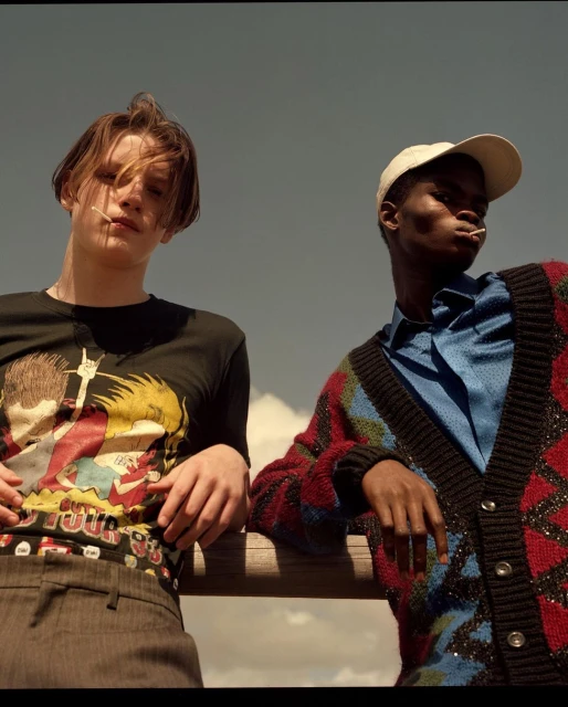 Two young adults pose outdoors on a sunny day. The individual on the left wears a graphic tee, while the person on the right sports a colorful cardigan.