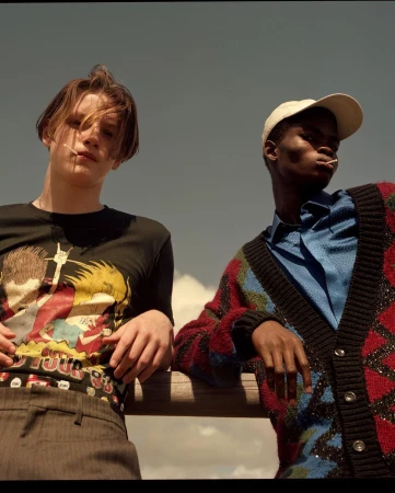 Two young adults pose outdoors on a sunny day. The individual on the left wears a graphic tee, while the person on the right sports a colorful cardigan.