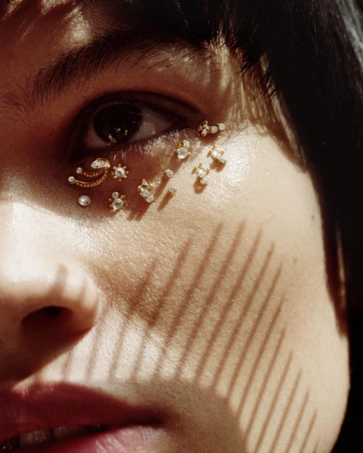 Close-up of an adult woman's eye, adorned with small, ornate gold and jeweled decorations, with strong shadow pattern across the face.
