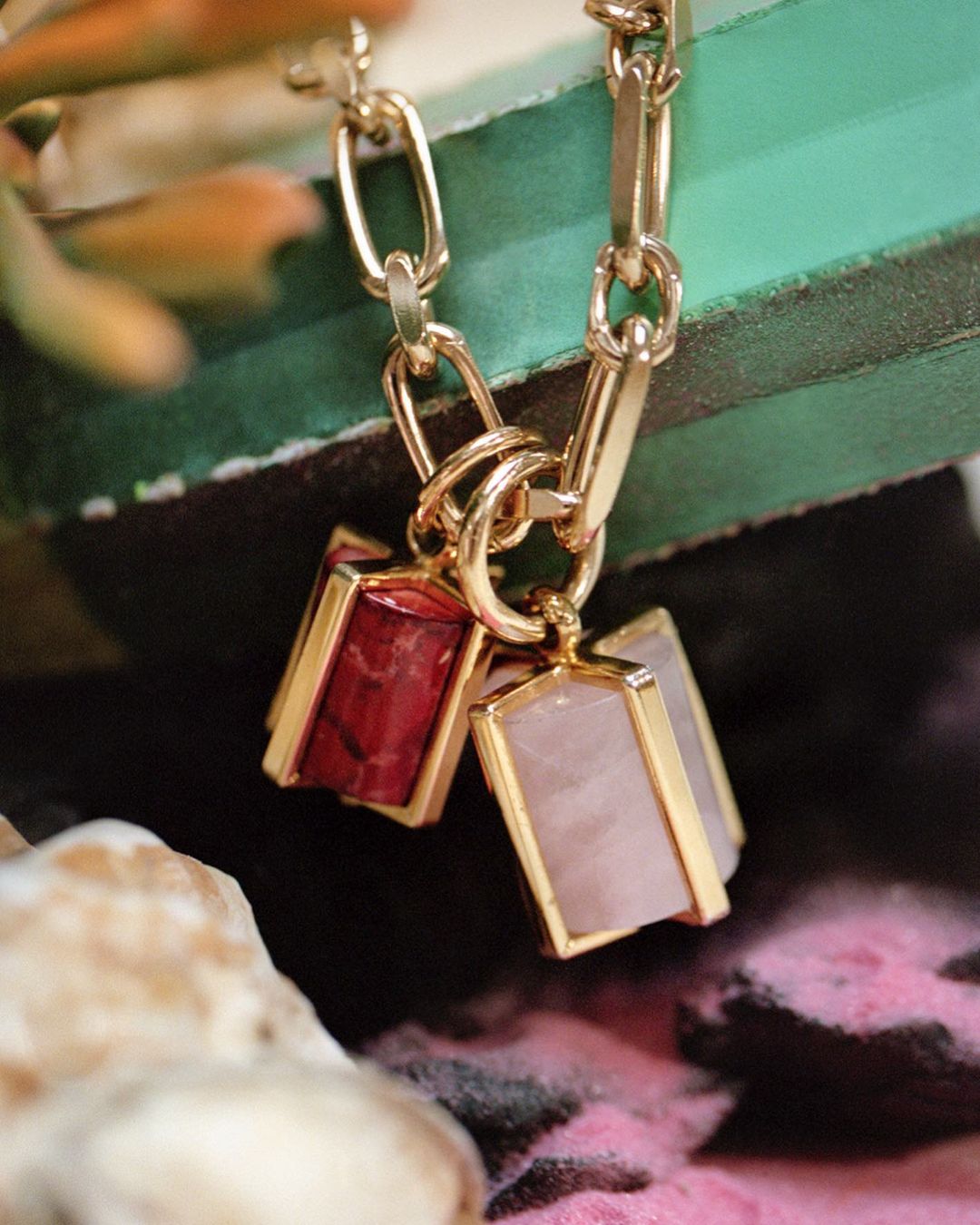 Close-up studio shot of a gold chain necklace with two charm pendants: one red, one pink, against a pink and green background.