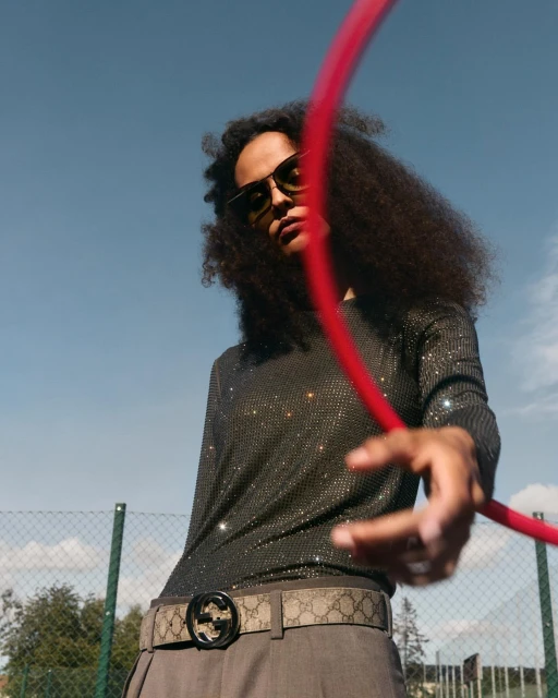 Adult posing with a red hoop outdoors. The model wears sunglasses, a long-sleeved shirt, grey pants, and Gucci belt against a blue sky.