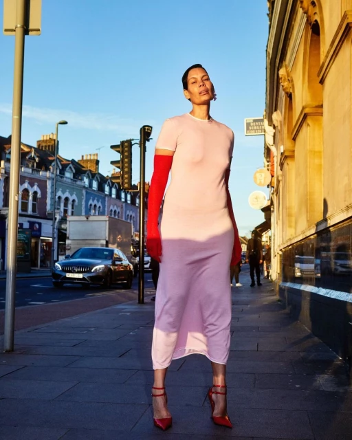 Fashion shot of an adult woman in a pink dress and red gloves standing on a sidewalk with buildings and a car in the background.