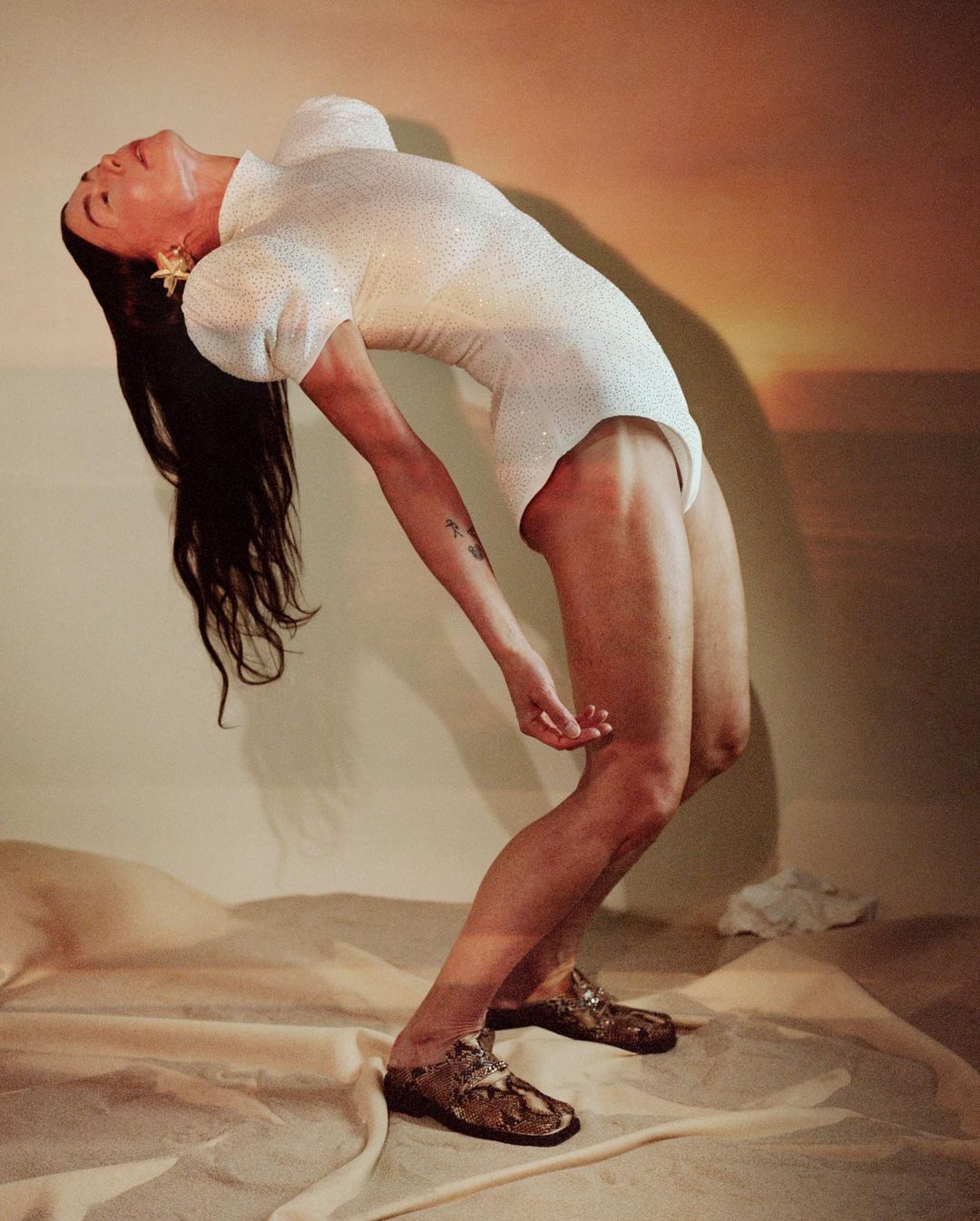 A woman in a white bodysuit and metallic shoes arches her back in a studio setting with warm-toned background lighting.
