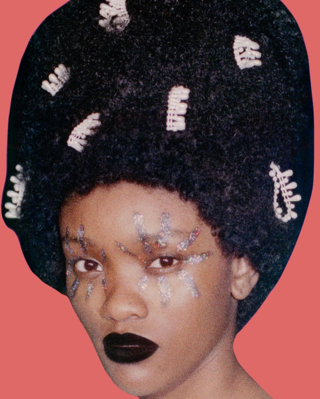 Portrait of a woman with dark lipstick and elaborate glitter eye makeup, her afro decorated with white hair accessories against a coral background.