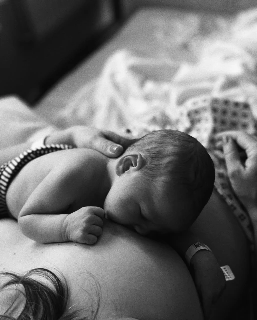 Black and white close-up of a newborn infant nursing at their mother's breast, with the mother's hand gently holding the baby's head.