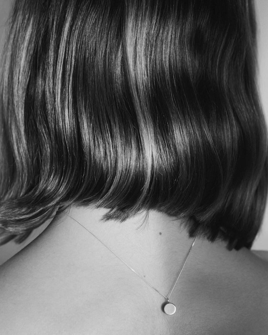 Black and white close-up shot of an adult woman's back of head, bob haircut, and a simple pendant necklace on a delicate chain.