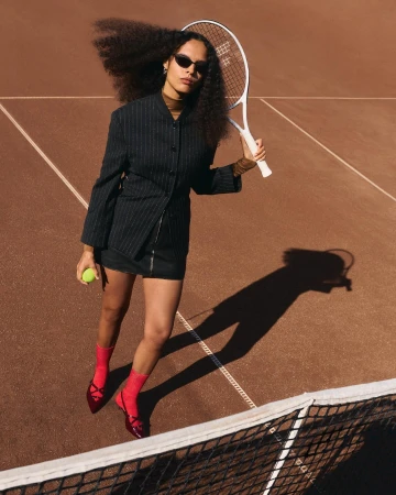 Woman in dark pinstripe jacket holding tennis racket and ball on a clay tennis court