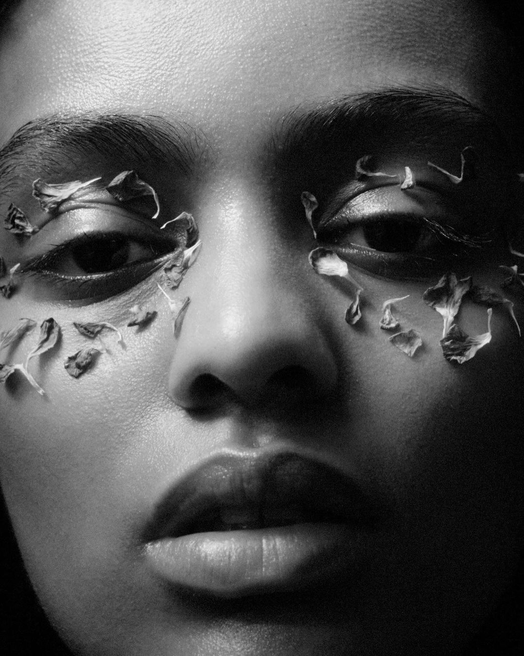 Close-up portrait of a woman in black and white, with delicate flower petals arranged around her eyes as artistic decoration.
