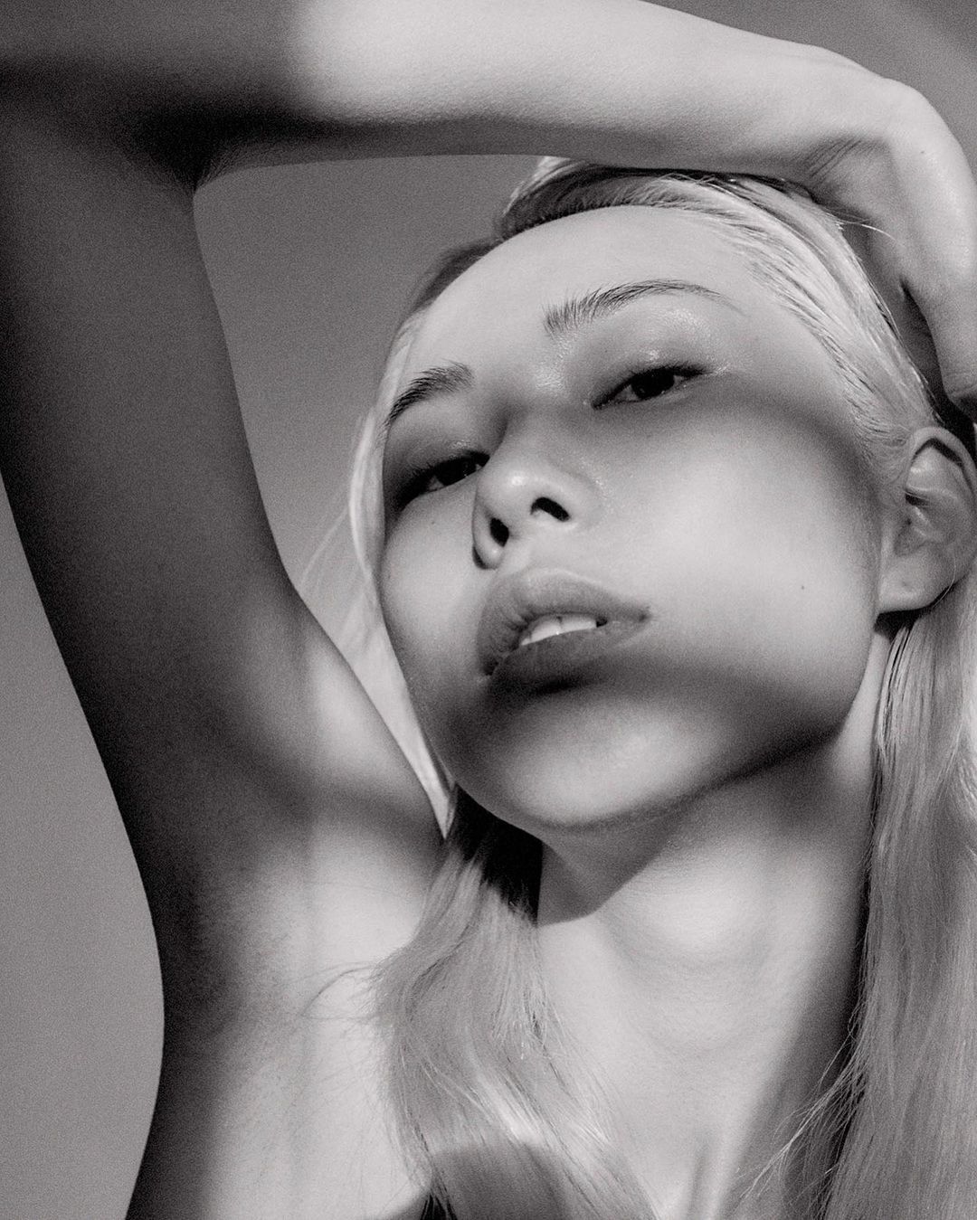 Monochrome portrait of a blonde woman with hand on head, eye-level angle, soft lighting, and neutral background in a beauty shot.