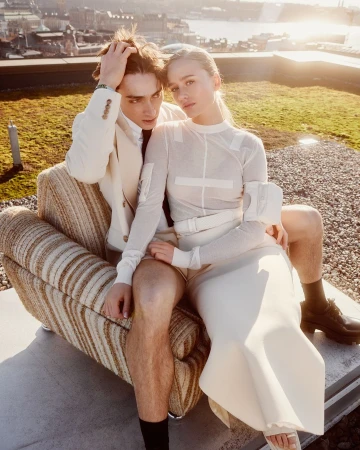A young couple seated together in an armchair on a sunny rooftop overlooking a city skyline at sunset.