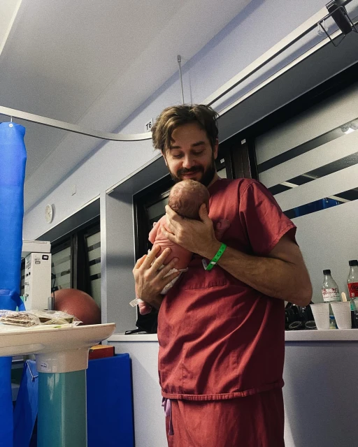 A man in scrubs holds a newborn baby indoors with a hospital setting visible in the background.