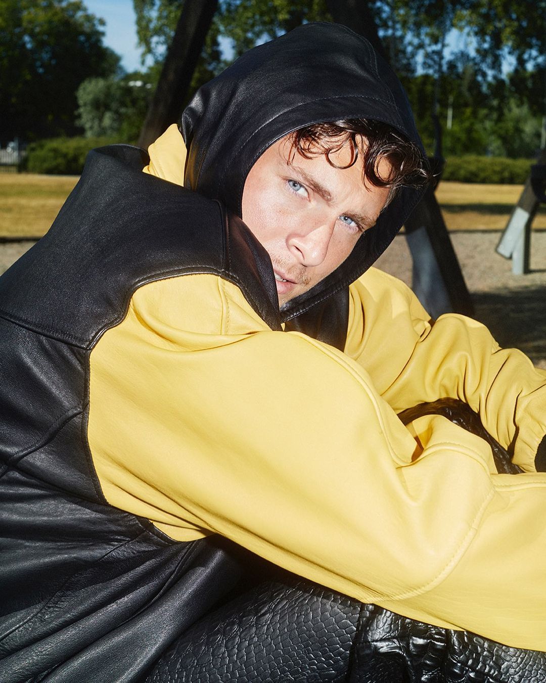 Young adult male with blue eyes wearing a black and yellow hooded leather jacket sitting on a playground swing outdoors in daytime.