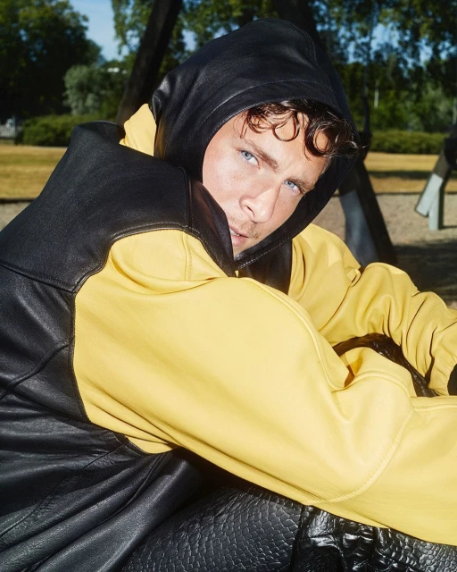 Young adult male with blue eyes wearing a black and yellow hooded leather jacket sitting on a playground swing outdoors in daytime.