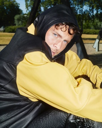 Young adult male with blue eyes wearing a black and yellow hooded leather jacket sitting on a playground swing outdoors in daytime.