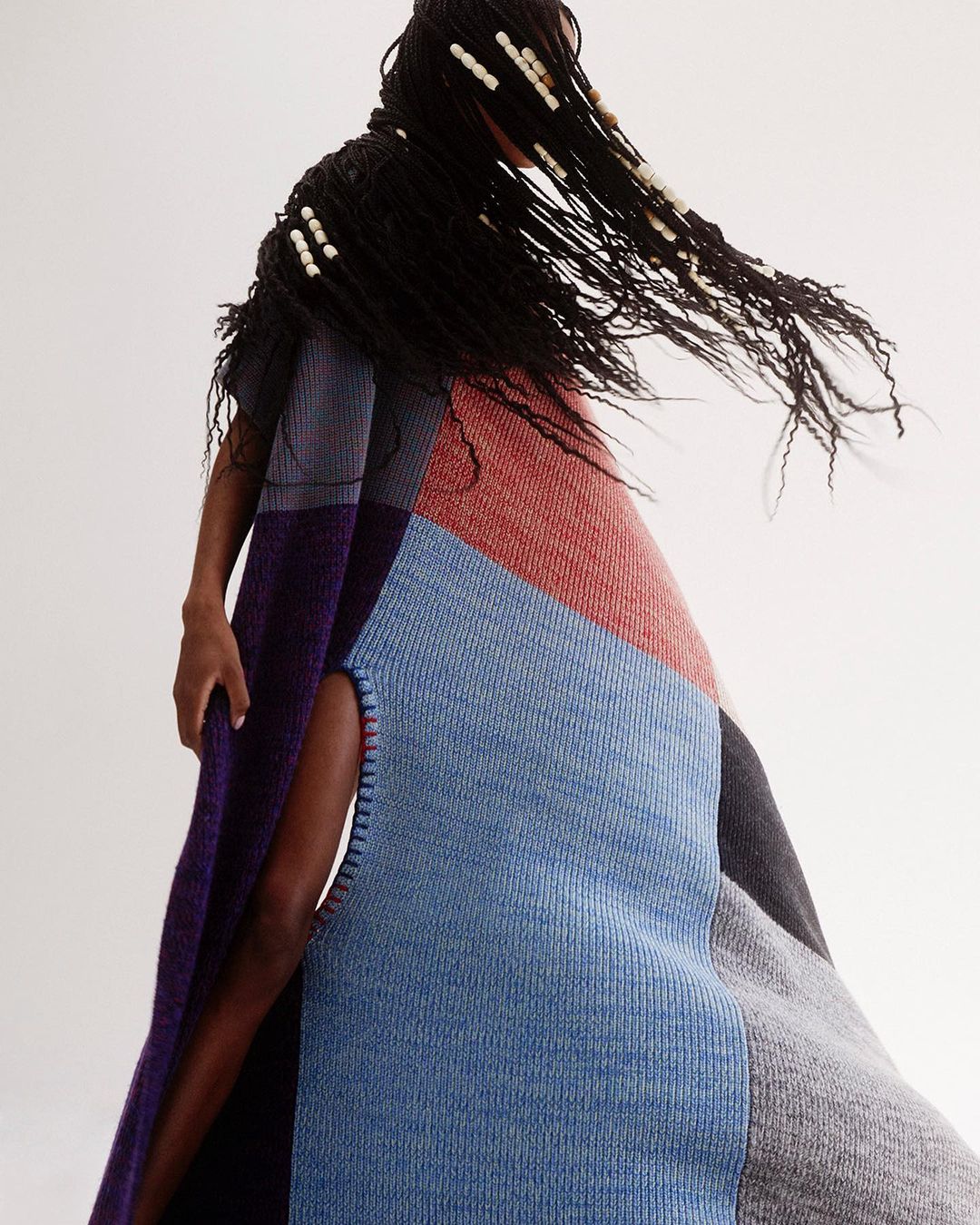 Fashionable person in a stylish patchwork woolen poncho with dark hair, adorned with beads, poses against a light neutral backdrop.