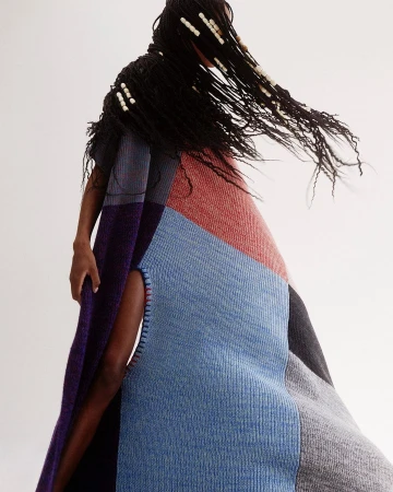 Fashionable person in a stylish patchwork woolen poncho with dark hair, adorned with beads, poses against a light neutral backdrop.