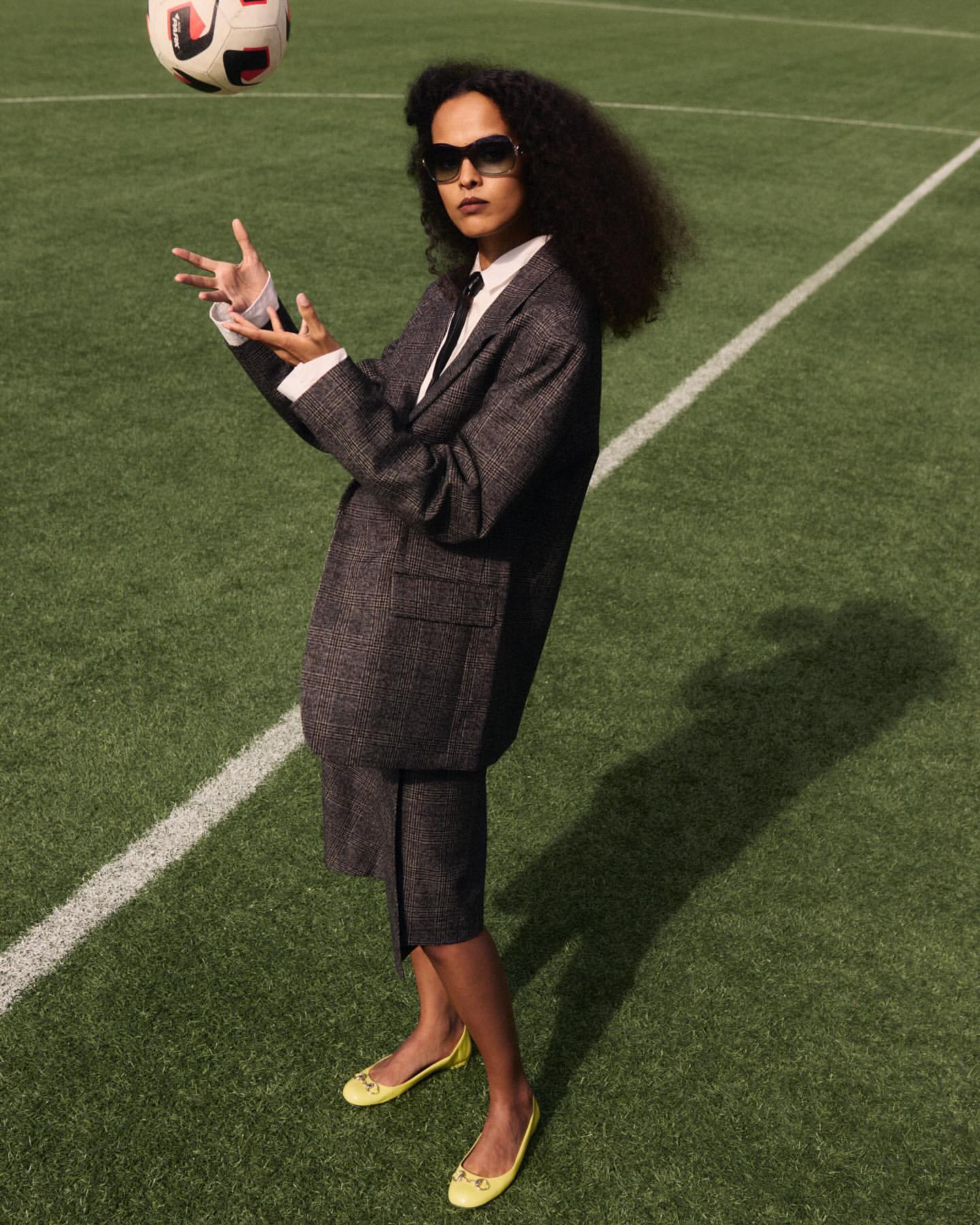 A woman in a suit and flats poses on a soccer field, juggling a soccer ball with her hands in outdoor daylight setting.