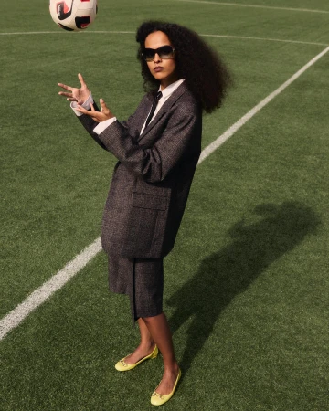 A woman in a suit and flats poses on a soccer field, juggling a soccer ball with her hands in outdoor daylight setting.
