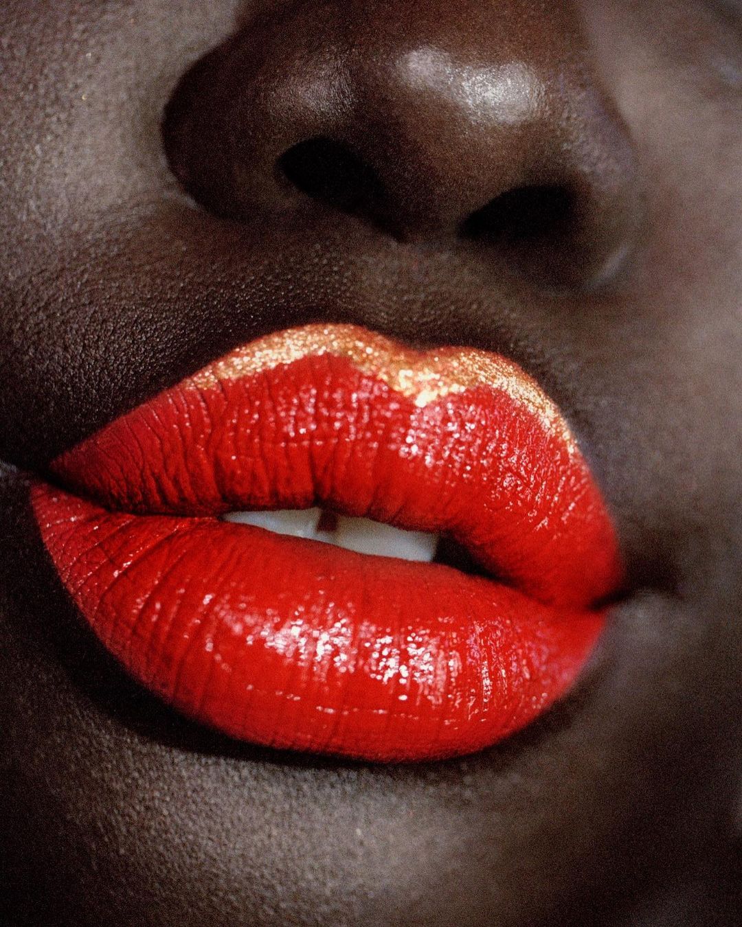 Close-up of a woman's face featuring dark skin, full red lips with gold glitter accents, and visible teeth, shot in a beauty style.