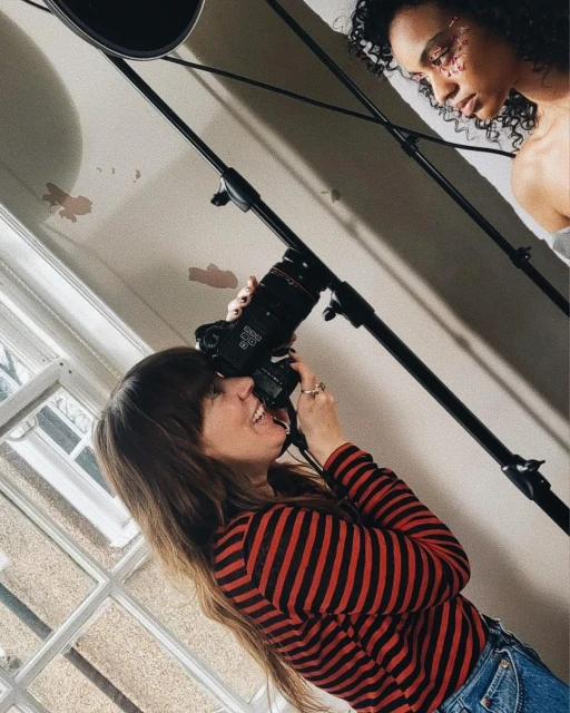 Female photographer captures a model in a studio, utilizing a DSLR camera on a stand. A window provides natural light in the space.