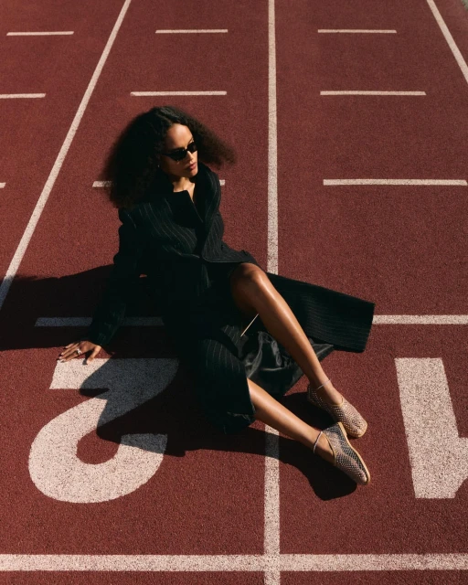 An adult female in a dark coat and sunglasses sits on a red running track with white lines, bathed in sunlight, fashion-forward style.