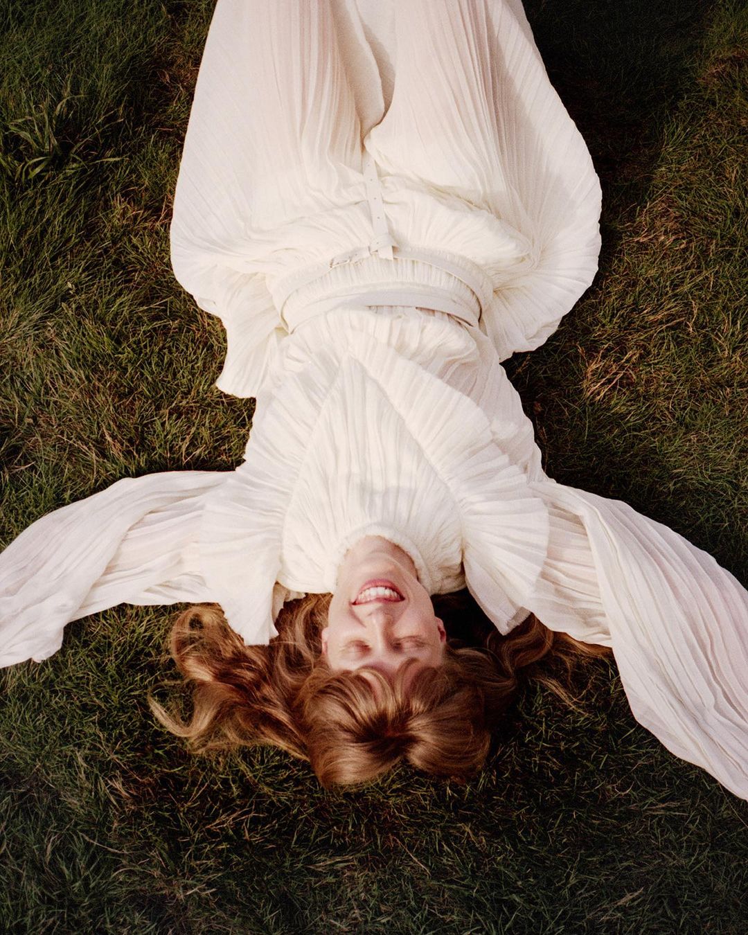 Woman in a flowing white dress reclines on green grass in a high-angle portrait, looking up with a smile. Her red hair is spread around her.