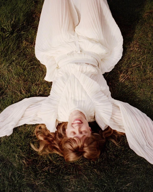 Woman in a flowing white dress reclines on green grass in a high-angle portrait, looking up with a smile. Her red hair is spread around her.