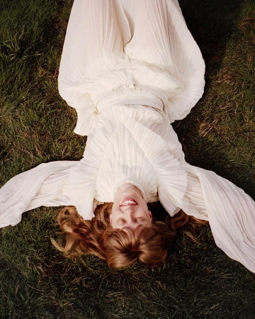 Woman in a flowing white dress reclines on green grass in a high-angle portrait, looking up with a smile. Her red hair is spread around her.