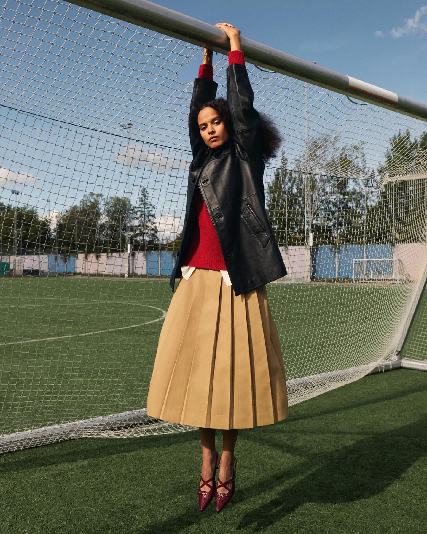 A woman with curly hair poses suspended from a metal bar, wearing a black jacket, red sweater, tan pleated skirt, and red heels on a green field.