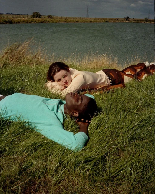 Two people, one Black and one white, lie side-by-side in a grassy field near a lake under a cloudy sky, creating a peaceful outdoor scene.